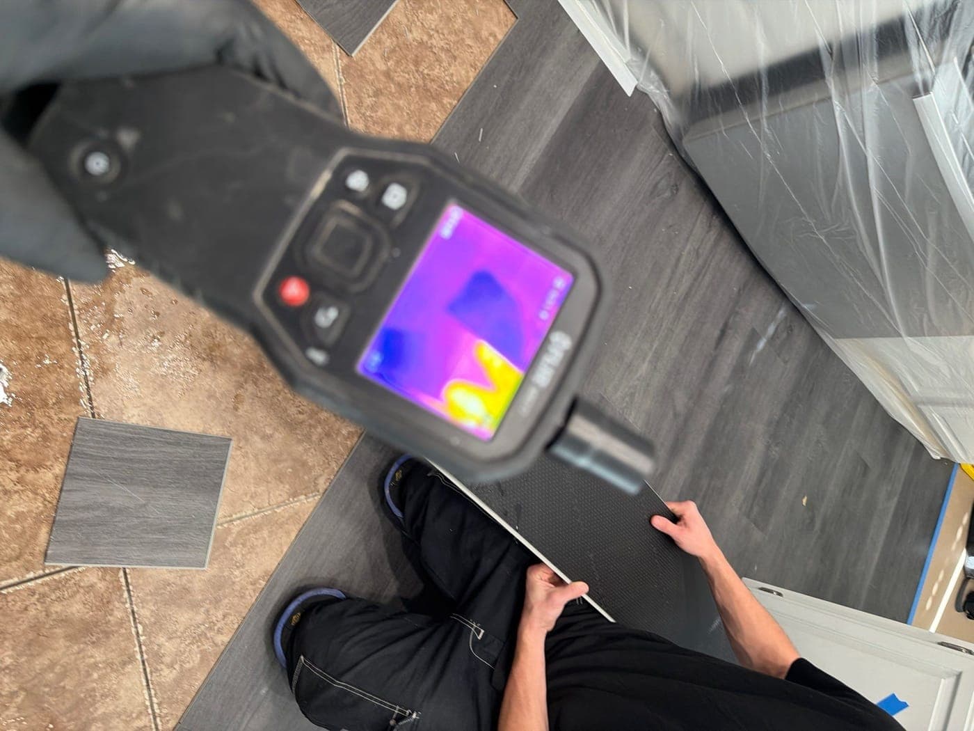 A thermal camera screen showing a cold blue signature where moisture sits behind the surface, with a technician working in the room beyond